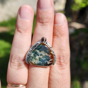 May include: A triangular ring featuring a large, polished gemstone with green, brown, and white patterns. The stone is set in a silver-toned metal frame with a small, copper-colored accent at the top. The ring is worn on a finger.