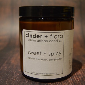 May include: A brown glass candle jar with a white label that reads "cinder + flora clean artisan candles sweet + spicy coconut, mandarin, chili pepper made by hand in Carlsbad, CA batch 12 6001".