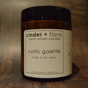 May include: A brown glass candle jar with a white label that reads "cinder + flora clean artisan candles rustic galette vanilla. butter. apple. made by hand in Carlsbad, CA".