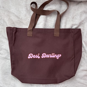 May include: A brown tote bag with brown handles. The bag features the text "Desi Darling" in a pink, cursive font. The bag is made of a sturdy material and is suitable for carrying various items.