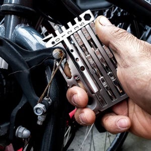 May include: A silver-toned multi-tool featuring a series of integrated metal bits and a ruler scale marked 0 through 5. The tool is constructed from flat metal plates with various cutouts and slots, designed for mechanical adjustments on a bicycle.