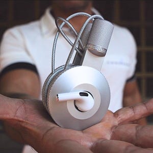 May include: A silver and grey headphone holder with a distinctive design. The holder has a circular base with a spiral pattern, a cylindrical top, and a cable that wraps around the design. A white earbud is inserted into the holder.