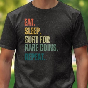 May include: A dark heather grey t-shirt with a vintage-style graphic that reads "Eat. Sleep. Sort for Rare Coins. Repeat."