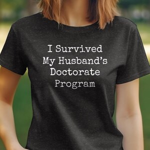 May include: A dark grey t-shirt with white text that reads "I Survived My Husband's Doctorate Program".