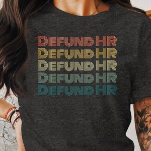 May include: A dark gray t-shirt with the text "DEFUND HR" repeated five times in a retro rainbow color scheme.