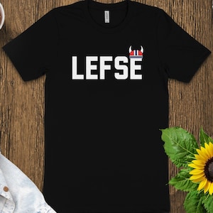 May include: Black t-shirt with the word "LEFSE" in white block letters. A small graphic of a Viking helmet in red, white, and blue is above the "E". The shirt is displayed on a wooden surface with a denim jacket, a succulent plant, and a sunflower.