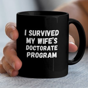 Può includere: Tazza da caffè nera con testo bianco che dice "Sono sopravvissuto al dottorato di mia moglie".