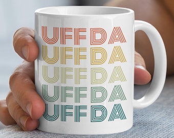Op de afbeelding: Witte keramische mok met het woord "UFFDA" vijf keer herhaald in een regenboogkleurige gradiënt. Elk woord is gemaakt van gestapelde lijnen.