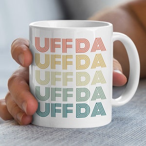 Peut inclure: Une tasse en céramique blanche avec le mot "UFFDA" répété cinq fois dans un dégradé de couleurs arc-en-ciel. Chaque mot est composé de lignes empilées.