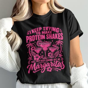 May include: A black short-sleeve t-shirt featuring a vibrant pink graphic print. The design displays three margarita glasses surrounded by palm trees and hibiscus flowers, with the text: I KEEP TRYING TO MAKE PROTEIN SHAKES BUT THEY KEEP COMING OUT AS MARGARITAS.