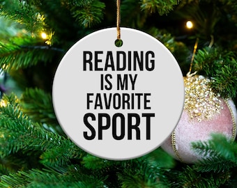 May include: A round, white Christmas ornament with the text "READING IS MY FAVORITE SPORT" in black. The ornament hangs from a gold string against a backdrop of a green Christmas tree and a pink and gold ornament.