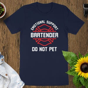 May include: Navy blue t-shirt with the text "EMOTIONAL SUPPORT BARTENDER DO NOT PET" in white and red. The design includes a red fire department emblem. The shirt is displayed on a wooden surface.