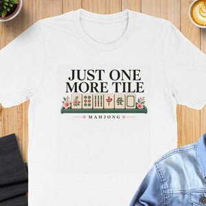 May include: A white short-sleeved t-shirt featuring a graphic print with the text JUST ONE MORE TILE and MAHJONG. The design includes a row of six mahjong tiles on a green rack, decorated with pink cherry blossom flowers on both ends.