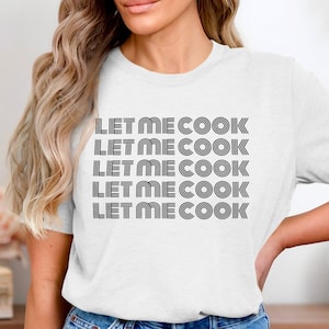 May include: White t-shirt with the text "LET ME COOK" in a repeating pattern. The text is in a bold, retro font. The shirt is a casual, everyday wear item.
