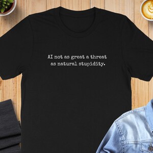 May include: Black t-shirt with the white text "AI not as great a threat as natural stupidity." The shirt is displayed on a wooden surface with a denim jacket, a cup of coffee, and a small plant.