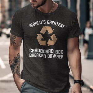May include: Dark gray t-shirt with the text "WORLD'S GREATEST CARDBOARD BOX BREAKER DOWNER" in white. The shirt features a recycling symbol in a tan color. The shirt is short-sleeved.