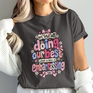 Puede incluir: Una camiseta gris carbón con un gráfico colorido que incluye el texto "we are all doing our best which is kind of embarrassing tbh". El diseño presenta patrones de cuadros rosas, azules y blancos, pequeñas flores y lazos de color rosa claro.