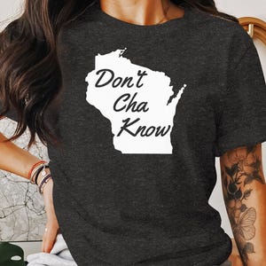 May include: Dark gray t-shirt featuring a white outline of the state of Wisconsin with the words "Don't Cha Know" in a handwritten font. The shirt is made of a soft material and is suitable for casual wear.