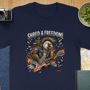May include: Navy blue t-shirt with a graphic of an eagle playing an American flag guitar, wearing a leather jacket. The text "SHRED & FREEDOM!" is above the eagle. The shirt is on a wooden surface with a camera and a bowl of kiwi.