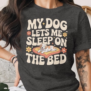 May include: Dark gray t-shirt with the text "MY DOG LETS ME SLEEP ON THE BED" in a retro font. The shirt features a graphic of a dog lying on a bed with flowers. A casual, comfortable tee.