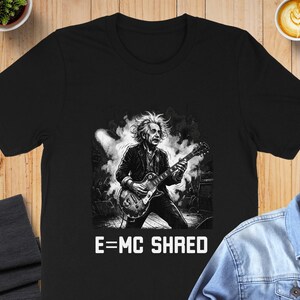 May include: Black t-shirt featuring a black and white illustration of Albert Einstein playing an electric guitar. The text "E=MC SHRED" is printed in white below the image. The shirt is displayed on a wooden surface.