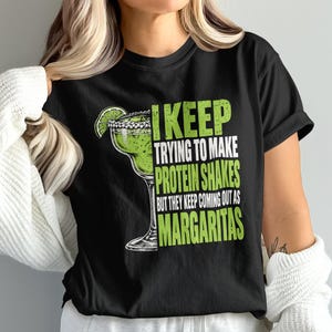 May include: A black t-shirt featuring a graphic of a green margarita in a glass with a lime wedge on the rim. The text reads: I KEEP TRYING TO MAKE PROTEIN SHAKES BUT THEY KEEP COMING OUT AS MARGARITAS. The lettering is in a bold, distressed green font.