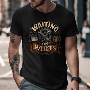 May include: A black short-sleeved t-shirt featuring a circular graphic with the text "WAITING ON PARTS". The design includes a wrench, a piston, a calendar, and a cardboard box, all rendered in shades of orange, blue, and grey on the chest.