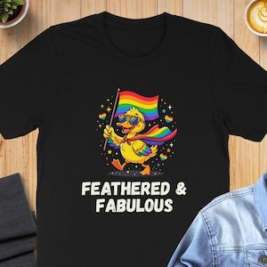 May include: A black t-shirt featuring a graphic of a yellow duck wearing sunglasses and a rainbow cape, holding a rainbow flag. The duck is surrounded by small colorful hearts and stars. Below the graphic, the text reads FEATHERED & FABULOUS.
