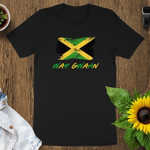 May include: Black t-shirt featuring the Jamaican flag design with a distressed look. The shirt has the words "WAH GWAAN" in green and yellow. The shirt is displayed on a wooden surface with a sunflower and a small plant.