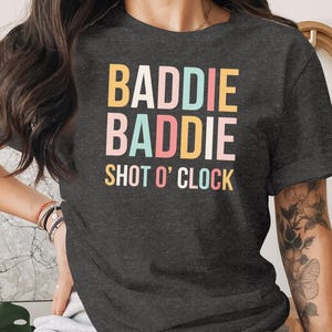 May include: Dark gray t-shirt with the words "BADIE BADIE SHOT O' CLOCK" in colorful block letters. The shirt is a casual style, suitable for everyday wear.