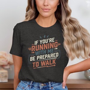May include: Dark gray t-shirt with the text "IF YOU'RE RUNNING WITH ME BE PREPARED TO WALK" in a retro design. The text is in shades of orange, teal, and white. The shirt is made of a soft material.