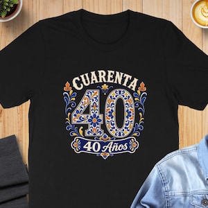 May include: A black short-sleeve t-shirt featuring a graphic print with the text CUARENTA and 40 Años. The design includes the number 40 decorated with blue, orange, and white floral patterns, surrounded by ornate blue and orange scrollwork.