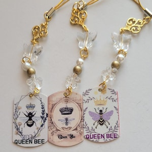 May include: Three decorative charms with gold-colored accents and the words "QUEEN BEE". Each charm features a tag with a bee illustration and a crown. The charms have clear butterfly and flower-shaped beads.