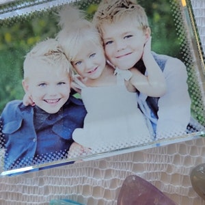 Puede incluir: Un marco de fotos de vidrio con una foto de tres niños sonriendo. El marco está sobre una superficie de crochet blanco con tres piedras de colores.