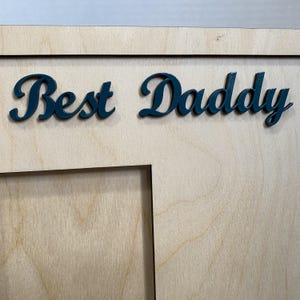 May include: Wooden picture frame with the words "Best Daddy" in teal script. The frame has a natural wood grain finish and a rectangular opening for a photograph.