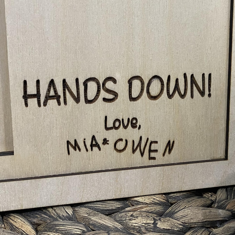 May include: A wooden sign with the phrase "HANDS DOWN!" in bold, dark letters. Below, the words "Love, MIA & OWEN" are written in a smaller, handwritten style. The sign has a light brown color.