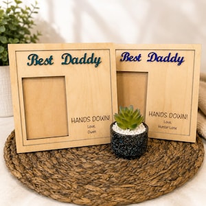 May include: Two wooden picture frames with the words "Best Daddy" in blue script. Each frame has a rectangular opening and the text "HANDS DOWN!" with a name below. A small succulent in a speckled pot sits between the frames.