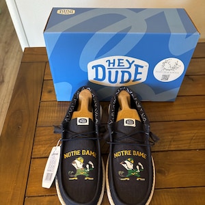 Peut inclure: Une paire de chaussures en toile noire avec une semelle blanche. Les chaussures présentent un logo Notre Dame et le texte "Notre Dame" sur elles. Les chaussures sont posées sur une surface en bois. Les chaussures sont dans une boîte sur laquelle est inscrit "Hey Dude".