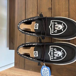 Pode incluir: Um par de sapatos de lona pretos com solas brancas, com o logotipo dos Raiders. Os sapatos têm um design com atacadores e um interior estampado. Uma etiqueta azul e branca com "HEY DUDE" está anexada.