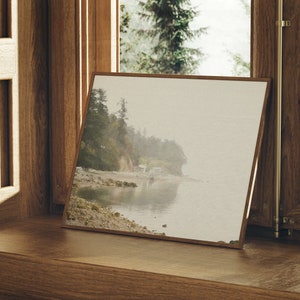 K&ouml;nnte beinhalten: Ein gerahmtes Foto einer nebeligen Strandszene mit felsiger K&uuml;ste und ruhigem Wasser. Das Foto ist in einem Holzrahmen und liegt auf einer Holzfensterbank.