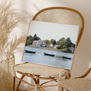 May include: A framed watercolor painting of a lake scene with three boats and a small town in the background. The painting is in a light wood frame and is sitting on a wicker chair.