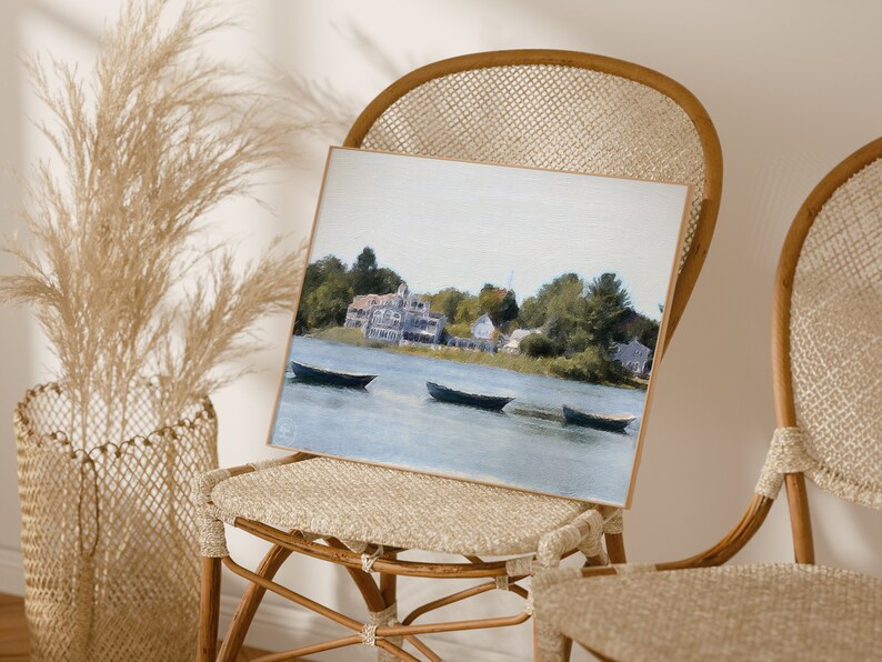 May include: A framed watercolor painting of a lake scene with three boats and a small town in the background. The painting is in a light wood frame and is sitting on a wicker chair.