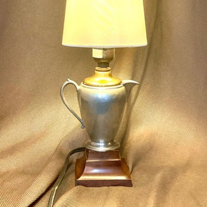 May include: A vintage-style table lamp features a pewter creamer base and a creamy off-white shade. The lamp's base is a pewter creamer with a classic shape and a dark brown wooden base. The lampshade is a soft, off-white fabric with subtle texture. The lamp is illuminated, casting a warm glow. This unique lamp would be a stylish addition to any home décor.