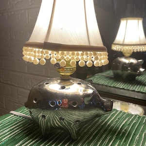 May include: Close-up view of a silver pig-shaped lamp base with small holes, topped with a white lampshade adorned with a beaded fringe. The lampshade is illuminated, casting a warm glow.  A similar lamp is reflected in a mirror-like surface behind it. The lamp rests on a green striped surface.