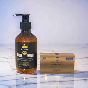 Pode incluir: Uma garrafa castanha de Oxylux Face & Body Moisturizer com um dispensador de bomba preto ao lado de uma caixa dourada de sabonete Oud For Him. A garrafa contém 300 ml de produto. A caixa de sabonete tem texto em inglês e francês.