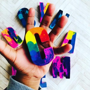 May include: A rainbow-colored crayon letter "C" is held in a hand, surrounded by other crayon letters. The letters are in various colors, including blue, pink, and yellow. The background is a light wood-grain surface.