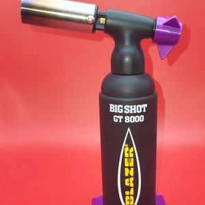 Puede incluir: Un encendedor de antorcha negro con una boquilla morada y una base hexagonal morada. La antorcha está etiquetada como "BIG SHOT GT 8000" y tiene la palabra "BLAZER" en letras amarillas.
