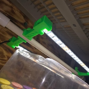 May include: Green plastic clips holding a white LED light strip to a white PVC pipe. The clips are fixed to a wooden surface. A clear plastic container with colourful circular objects is in the foreground.