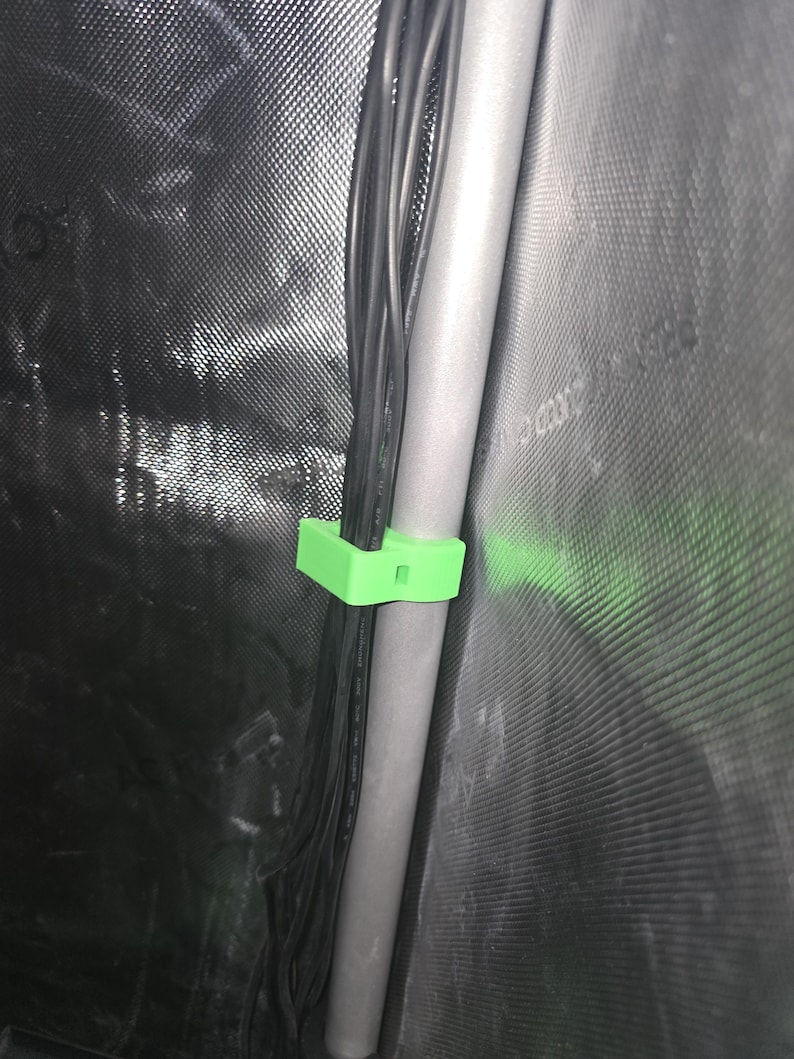 Puede incluir: Un clip de cable de pl&aacute;stico verde fijado a una tuber&iacute;a gris. El clip sujeta un haz de cables negros.