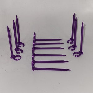 Puede incluir: Un juego de estacas de tienda de campaña de plástico morado con un lazo en la parte superior. Hay 10 estacas en total, con 5 estacas a cada lado de la imagen.